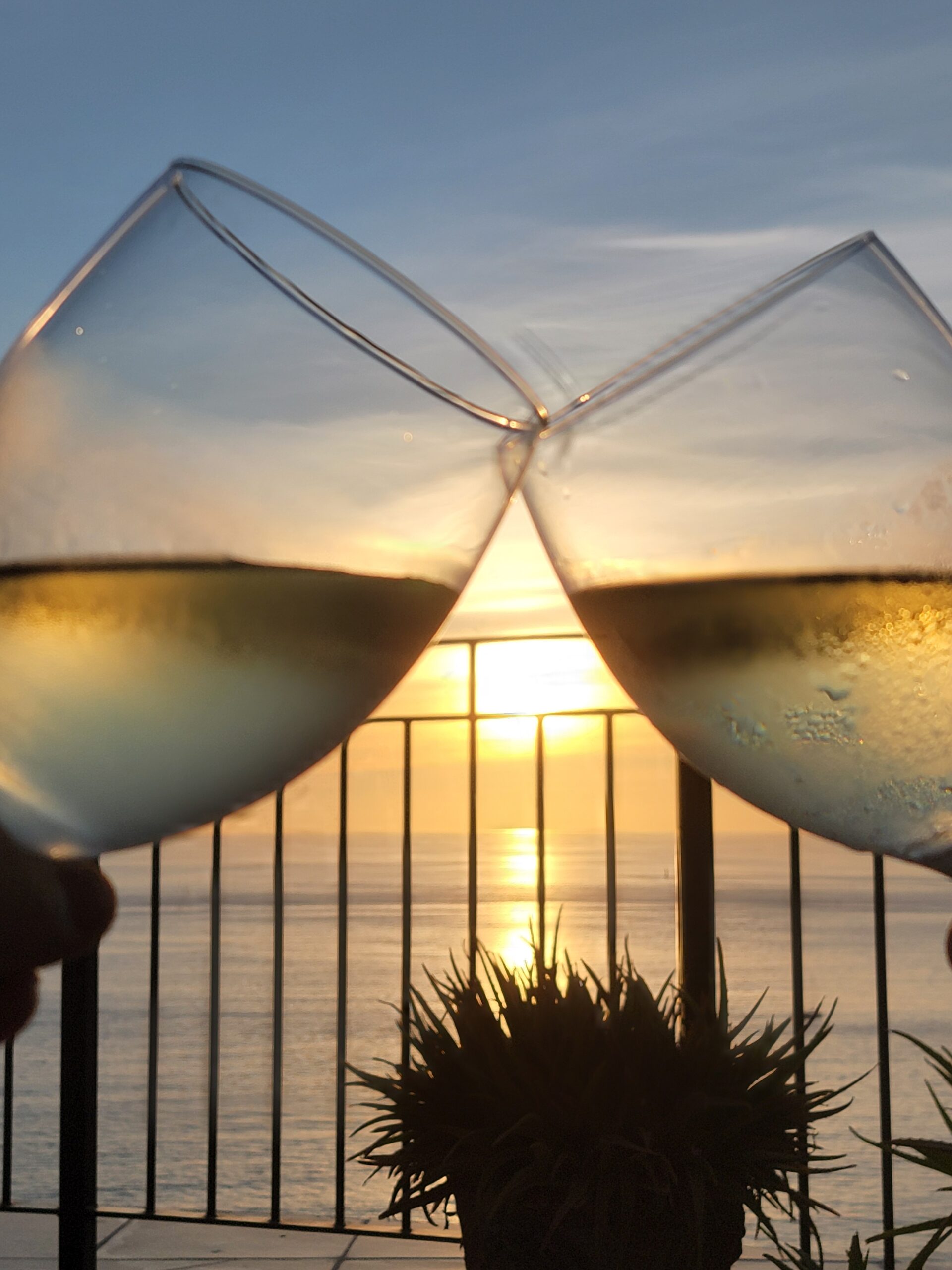 Two glasses of white wine clink together against a sunset over the ocean. The sun sits between the glasses, creating a beautiful effect. The glasses show condensation, indicating the wine is cool.